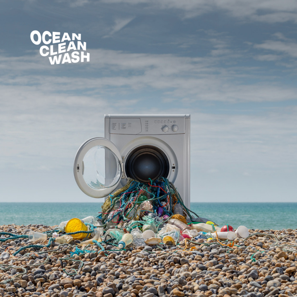Credit of the image: Oceancleanwash.org.  An images of a washing machine on the beach. Plastic and fishnet overflowing out of the washing machine onto the sandy beach.